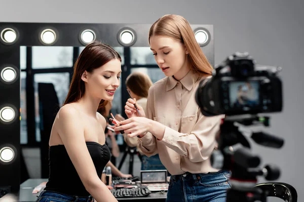 Selektiver Fokus des lächelnden Models in der Nähe des Make-up-Künstlers mit Lipgloss in der Nähe der Digitalkamera isoliert auf grau — Stockfoto