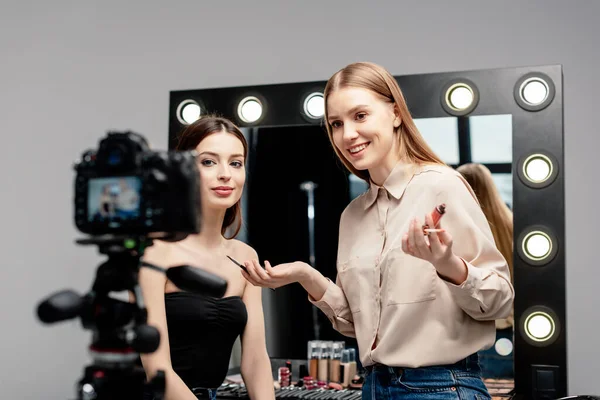 Foco seletivo do artista de maquiagem sorridente segurando o gloss perto do modelo e olhando para a câmera digital isolada no cinza — Fotografia de Stock