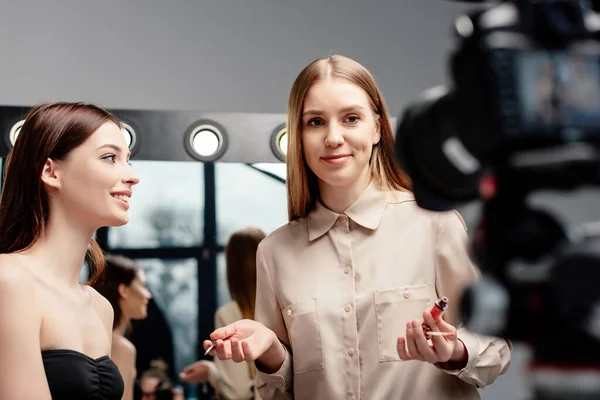 Selektiver Fokus des Maskenbildners, der Lipgloss in der Nähe des glücklichen Modells hält und die Digitalkamera isoliert auf grau betrachtet — Stockfoto