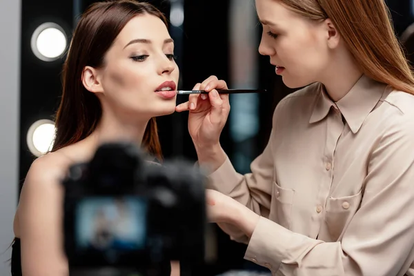 Selective focus of makeup artist applying lip gloss on lips of model near digital camera — Stock Photo