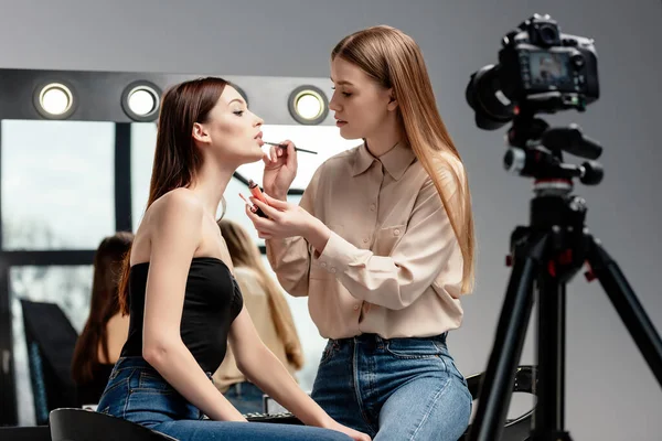 Mise au point sélective du maquilleur appliquant du brillant à lèvres sur les lèvres d'un beau modèle proche de l'appareil photo numérique isolé sur gris — Photo de stock