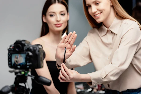 Selective focus of happy makeup artist holding cosmetic brush near beautiful model and digital camera isolated on grey — Stock Photo