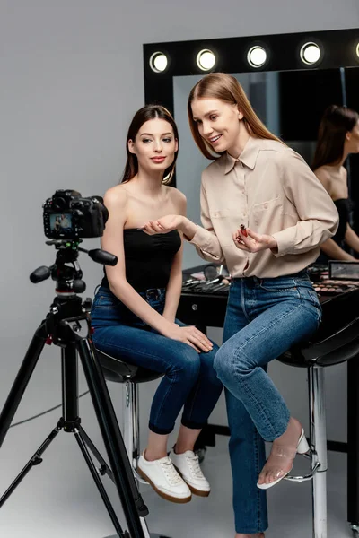 Selective focus of happy makeup artist holding lip gloss near beautiful model and digital camera on grey — Stock Photo