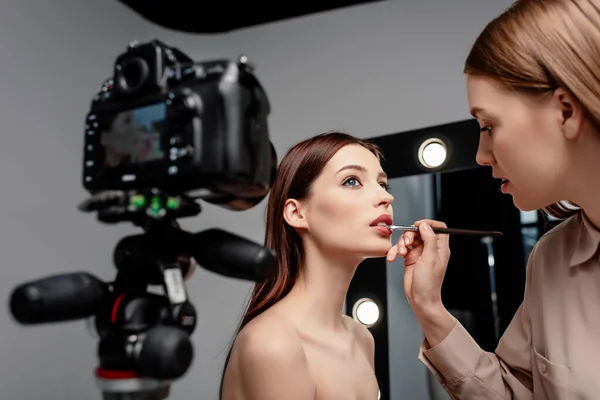 Foco seletivo do artista de maquilagem aplicando o lustro de lábio em lábios do modelo atraente perto da câmera digital no cinza — Fotografia de Stock