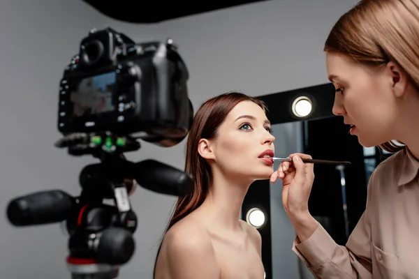 Messa a fuoco selettiva di trucco artista applicando lucidalabbra su labbra di modello attraente vicino a macchina fotografica digitale su grigio — Foto stock