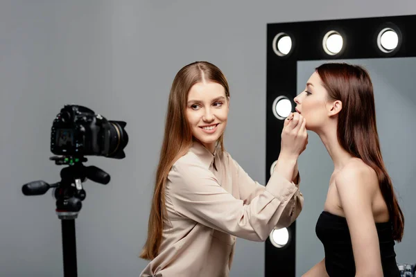 Foco seletivo de artista de maquiagem feliz aplicando brilho labial em lábios de modelo atraente e olhando para a câmera digital isolada em cinza — Fotografia de Stock