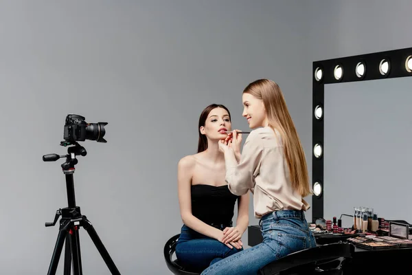 Artista de maquiagem alegre aplicando o lustro de lábio em lábios do modelo atraente e olhando para a câmera digital no cinza — Fotografia de Stock