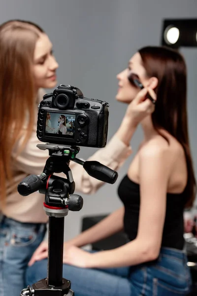 Mise au point sélective du trépied avec appareil photo numérique près du maquilleur heureux appliquant de la poudre visage sur un beau modèle — Photo de stock