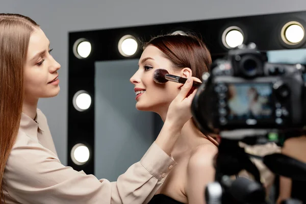 Selective focus of cheerful makeup artist applying face powder on beautiful model near digital camera — Stock Photo