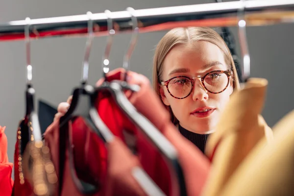 Selective focus of attractive stylist in glasses looking at fashionable clothing on hangers — Stock Photo