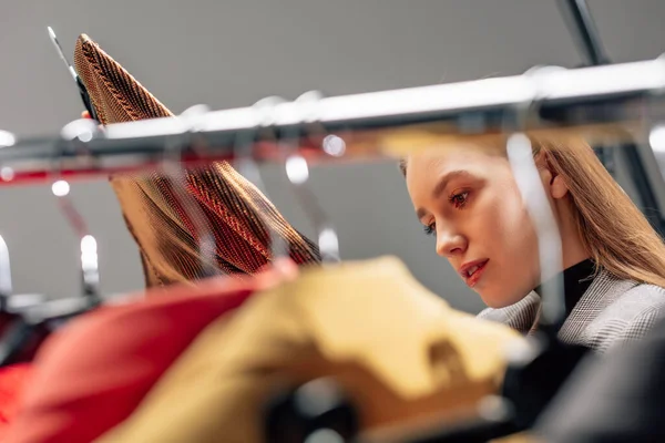 Selective focus of beautiful stylist looking at fashionable clothing on hangers — Stock Photo