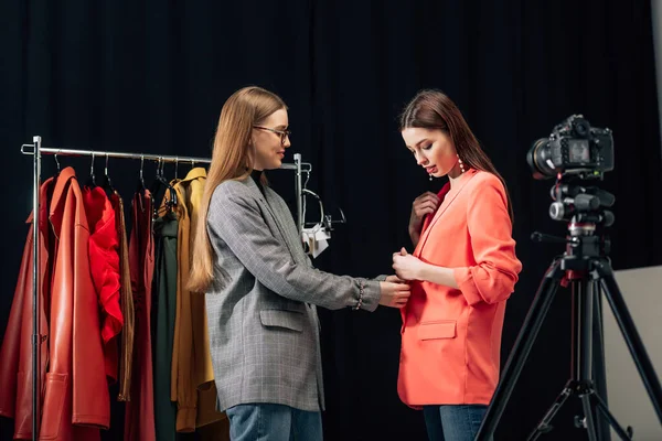 Избирательный фокус красивого стилиста в очках касающихся модной куртки на привлекательной модели рядом с цифровой камерой — стоковое фото