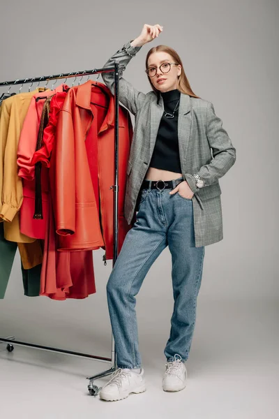 Estilista atractivo en gafas de pie con la mano en el bolsillo cerca de la ropa de moda en gris - foto de stock