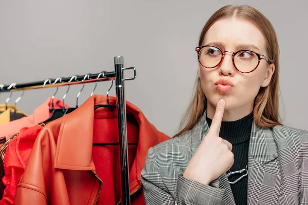 Pensive stylist in glasses touching face while thinking near trendy clothing isolated on grey — Stock Photo