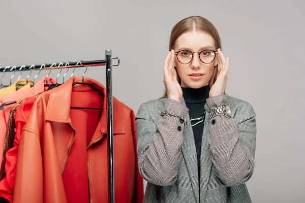 Junge Stylistin berührt Brille in der Nähe trendiger Kleidung isoliert auf grau — Stockfoto