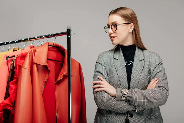 Giovane stilista in occhiali in piedi con le braccia incrociate vicino abbigliamento alla moda isolato su grigio — Foto stock