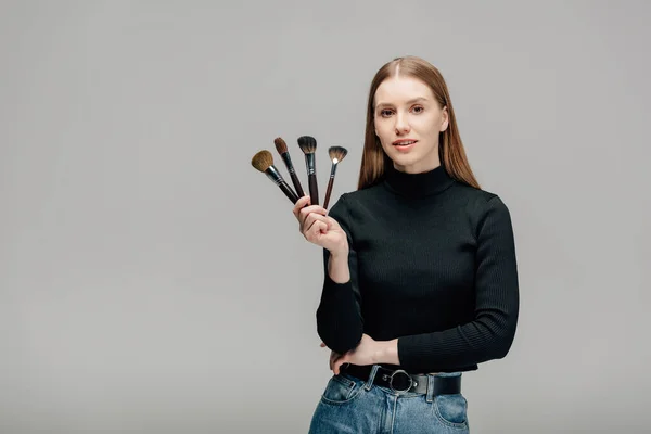 Attractive makeup artist holding makeup brush set isolated on grey — Stock Photo