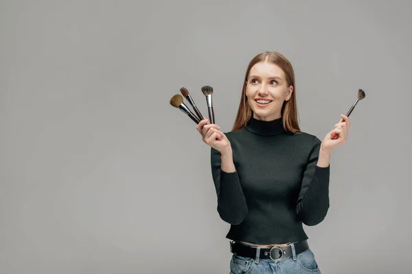 Cheerful makeup artist holding cosmetic brushes isolated on grey — Stock Photo