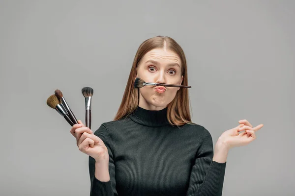 Artista de maquillaje divertido celebración de cepillos cosméticos aislados en gris - foto de stock
