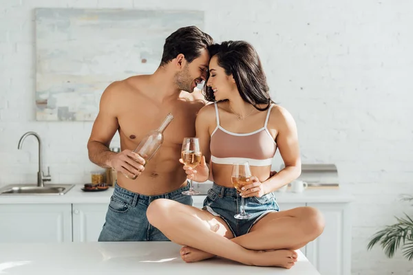Novio con botella de vino y novia con copas y piernas cruzadas sobre mesa en cocina - foto de stock