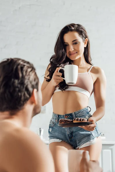 Enfoque selectivo de la mujer con taza de té y plato con rosquillas mirando novio - foto de stock