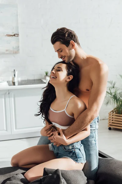 Hombre abrazando atractiva mujer feliz por detrás en el sofá en la cocina - foto de stock