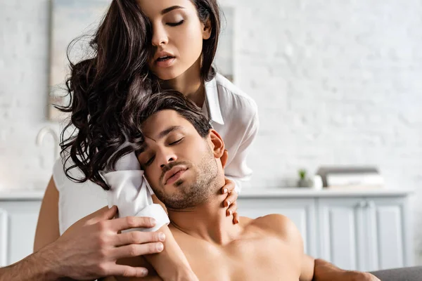 Mujer sexy en camisa blanca abrazando al hombre con los ojos cerrados en la cocina - foto de stock