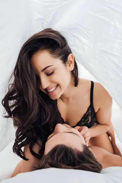 Sexy girl with closed eyes on boyfriend smiling under sheet on bed in bedroom — Stock Photo