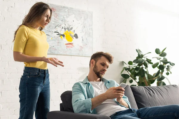 Angry woman standing near boyfriend with smartphone on couch — Stock Photo