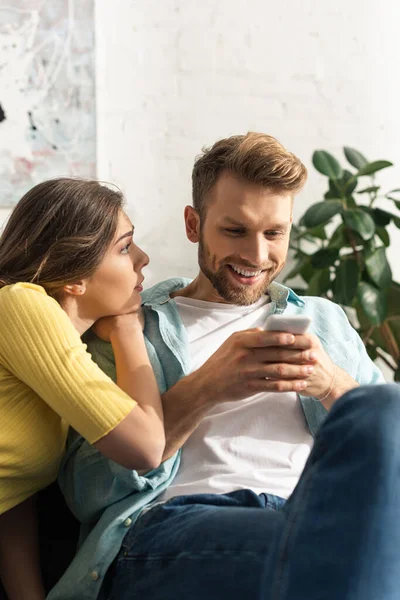 Donna guardando sorridente fidanzato utilizzando smartphone sul divano — Foto stock