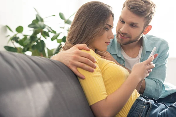 Sonriente hombre abrazando dependiente novia con teléfono inteligente en el sofá - foto de stock