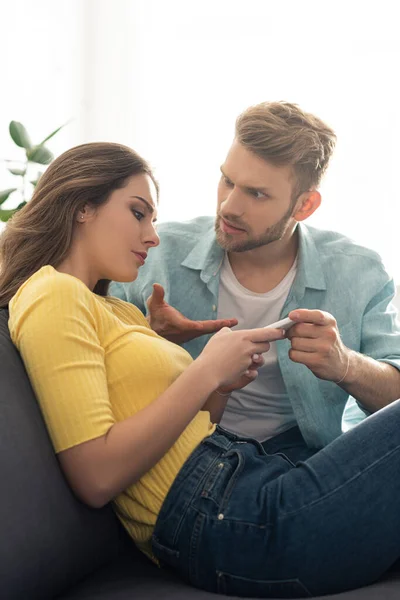 Hombre agresivo apuntando con la mano mientras su novia utiliza el teléfono inteligente en el sofá - foto de stock