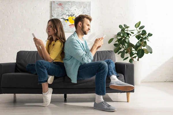 Vista lateral de la pareja usando teléfonos inteligentes mientras se sienta espalda con espalda en el sofá - foto de stock