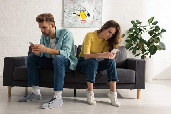 Pareja joven usando teléfonos inteligentes mientras está sentada en el sofá en casa - foto de stock