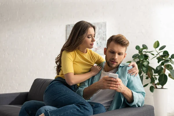 Hermosa mujer abrazando novio adicto con teléfono inteligente en el sofá - foto de stock