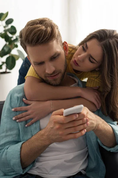 Attractive woman hugging handsome boyfriend using smartphone at home — Stock Photo