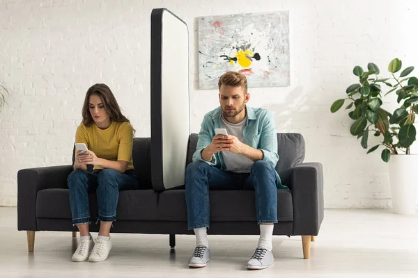 Pareja joven usando teléfonos inteligentes cerca de un enorme modelo de teléfono inteligente en el sofá - foto de stock