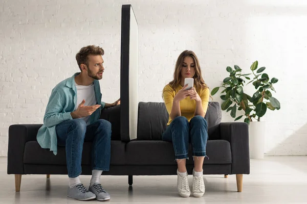 Confused man looking at girlfriend chatting near big model of smartphone on sofa — Stock Photo