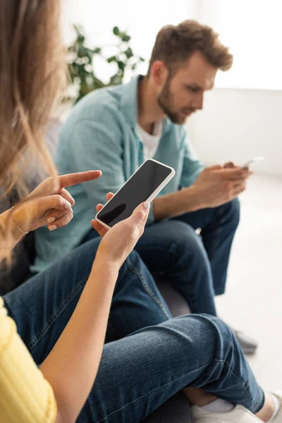 Mise au point sélective de la femme pointant du doigt le smartphone avec écran vide près du petit ami sur le canapé — Photo de stock