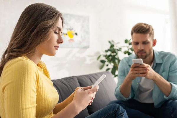 Enfoque selectivo de la mujer hermosa usando el teléfono inteligente cerca del hombre en el sofá en casa - foto de stock