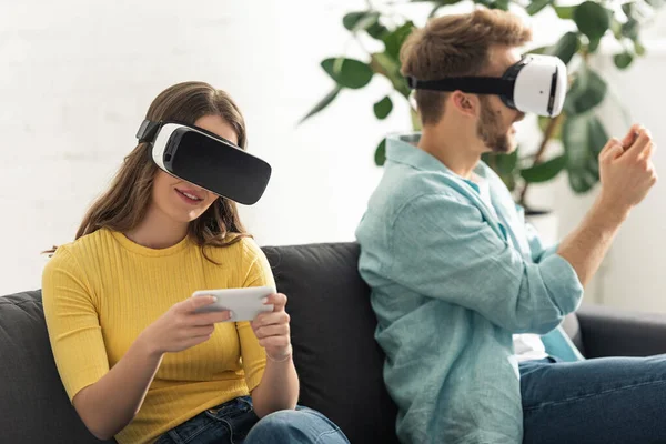 Selective focus of smiling girl in vr headset using smartphone near boyfriend on sofa in living room — Stock Photo