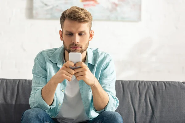 Hombre guapo usando teléfono inteligente mientras está sentado en el sofá en casa - foto de stock