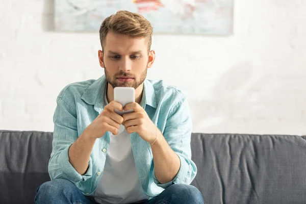 Bonito homem usando smartphone enquanto sentado no sofá em casa — Fotografia de Stock