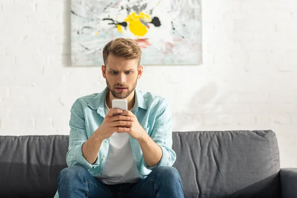 Uomo confuso utilizzando smartphone sul divano di casa — Foto stock