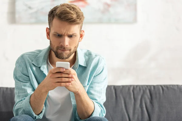 Uomo confuso guardando smartphone sul divano a casa — Foto stock
