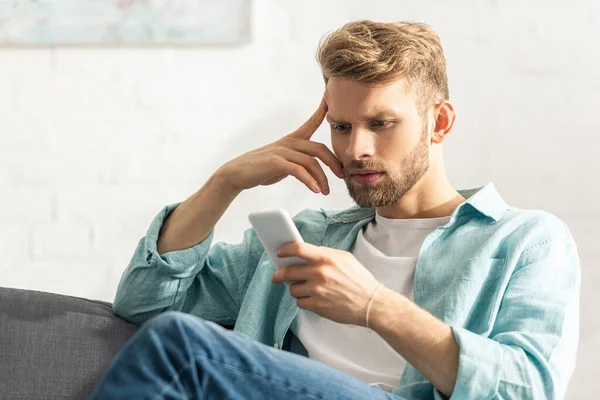 Pensive людина з пальцем біля чола, використовуючи смартфон на дивані — стокове фото