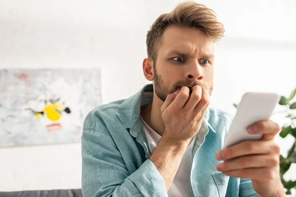 Selektiver Fokus eines verängstigten Mannes mit Hand in der Nähe des Mundes mittels Smartphone im Wohnzimmer — Stockfoto