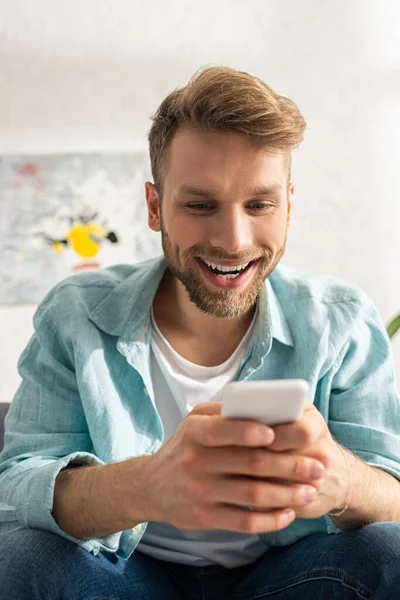 Enfoque selectivo del hombre adicto sonriente utilizando el teléfono inteligente en la sala de estar - foto de stock