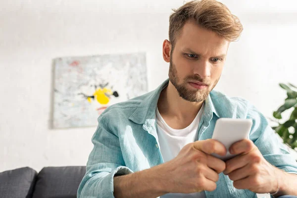 Selective focus of angry man using smartphone on sofa in living room — Stock Photo