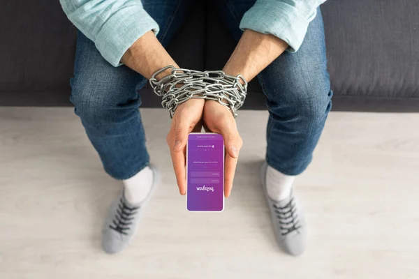 KYIV, UKRAINE - FEBRUARY 21, 2020: Top view of man with metal chain around hands holding smartphone with instagram app on couch — Stock Photo